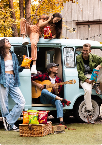 Four people smile while holding bags of chips and sitting around a blue van