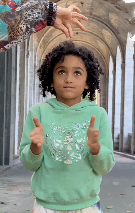 A boy in a green sweat shirt gives two thumbs up while looking up at someone's hand above his head