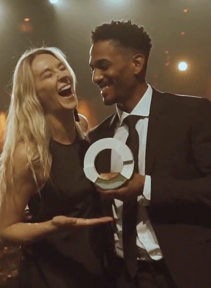 A man in a suit and a woman in a dress laugh while looking at each other